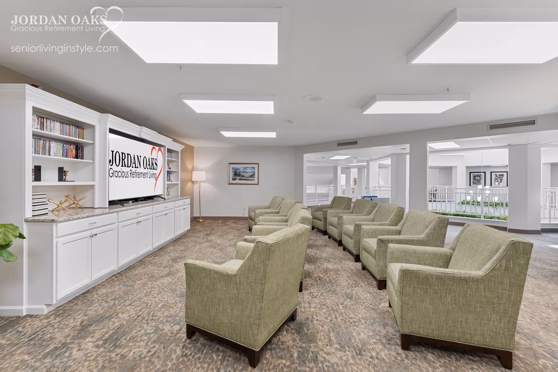 Spacious senior living lounge with rows of green armchairs facing a built-in TV and bookcases.