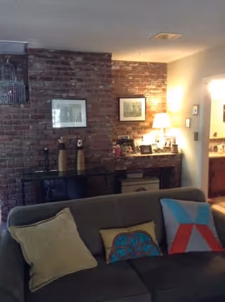 Cozy living room with a gray couch adorned with three decorative pillows in different colors and patterns. Behind the couch is a brick wall with two framed pictures and a console table holding decorative items. A lit table lamp and various framed photos are on a side table near an open doorway leading to a bathroom.
