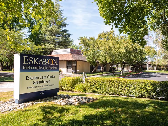 Exterior view of Eskaton Care Center Greenhaven showing the facility's sign in the foreground surrounded by greenery, trees, and a clear blue sky. The building is partially visible in the background with a driveway and landscaped bushes.