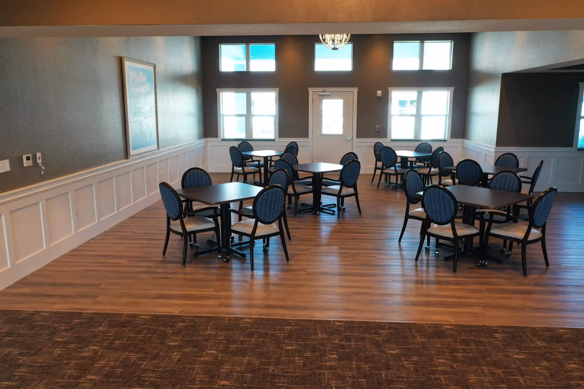 A dining room with several square tables, each surrounded by four blue cushioned chairs. The room has wooden flooring, white wainscoting on the walls, and multiple windows letting in natural light. There is a door at the far end of the room beneath a hanging light fixture.