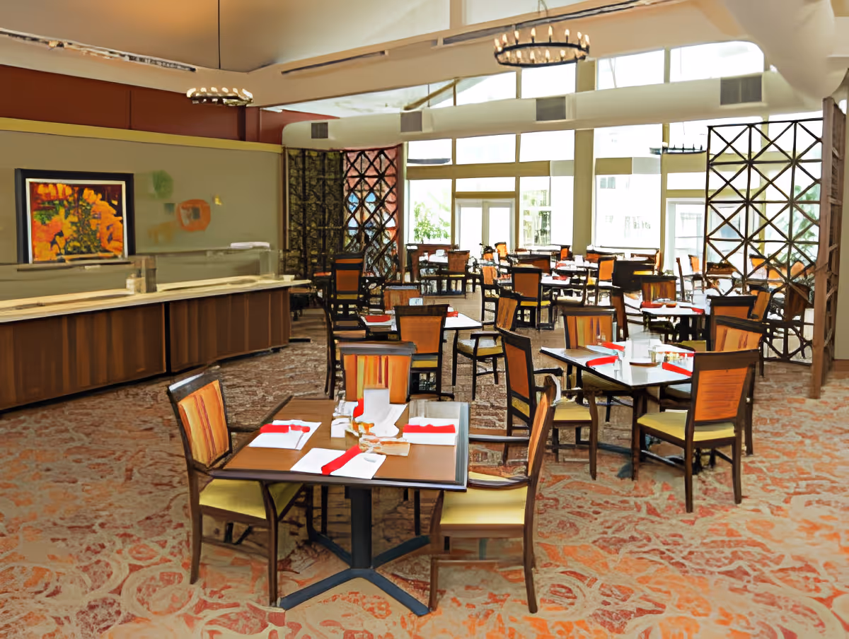 Bright dining room with multiple set tables and chairs, a buffet counter, and large windows.