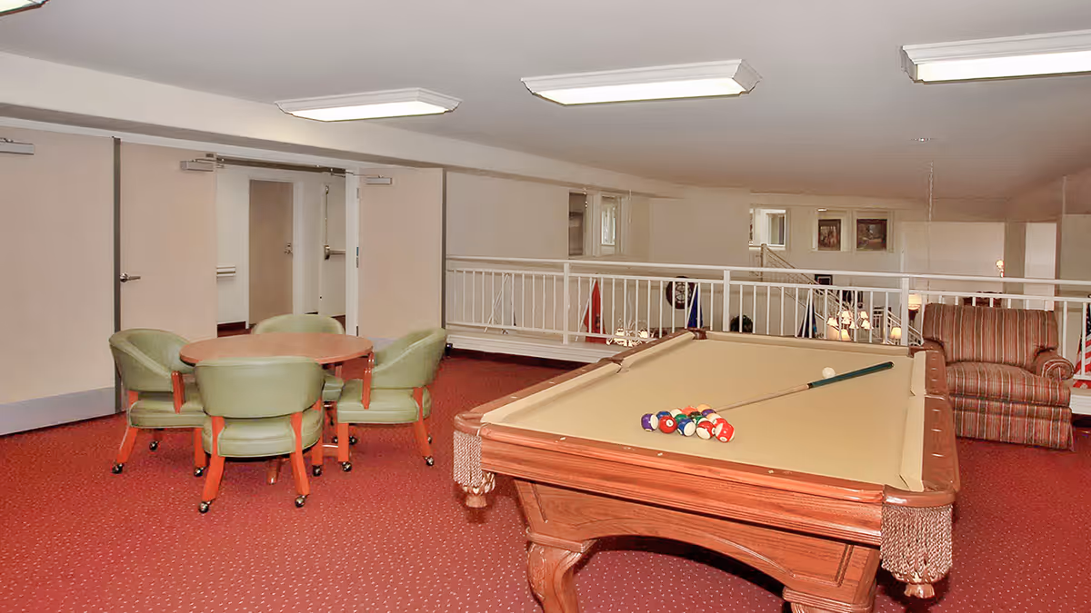 Interior room with a pool table set up for a game, a round table with four green chairs, and a striped armchair. The room has red carpet flooring, white walls, and ceiling lights. There is a white railing overlooking a lower level.