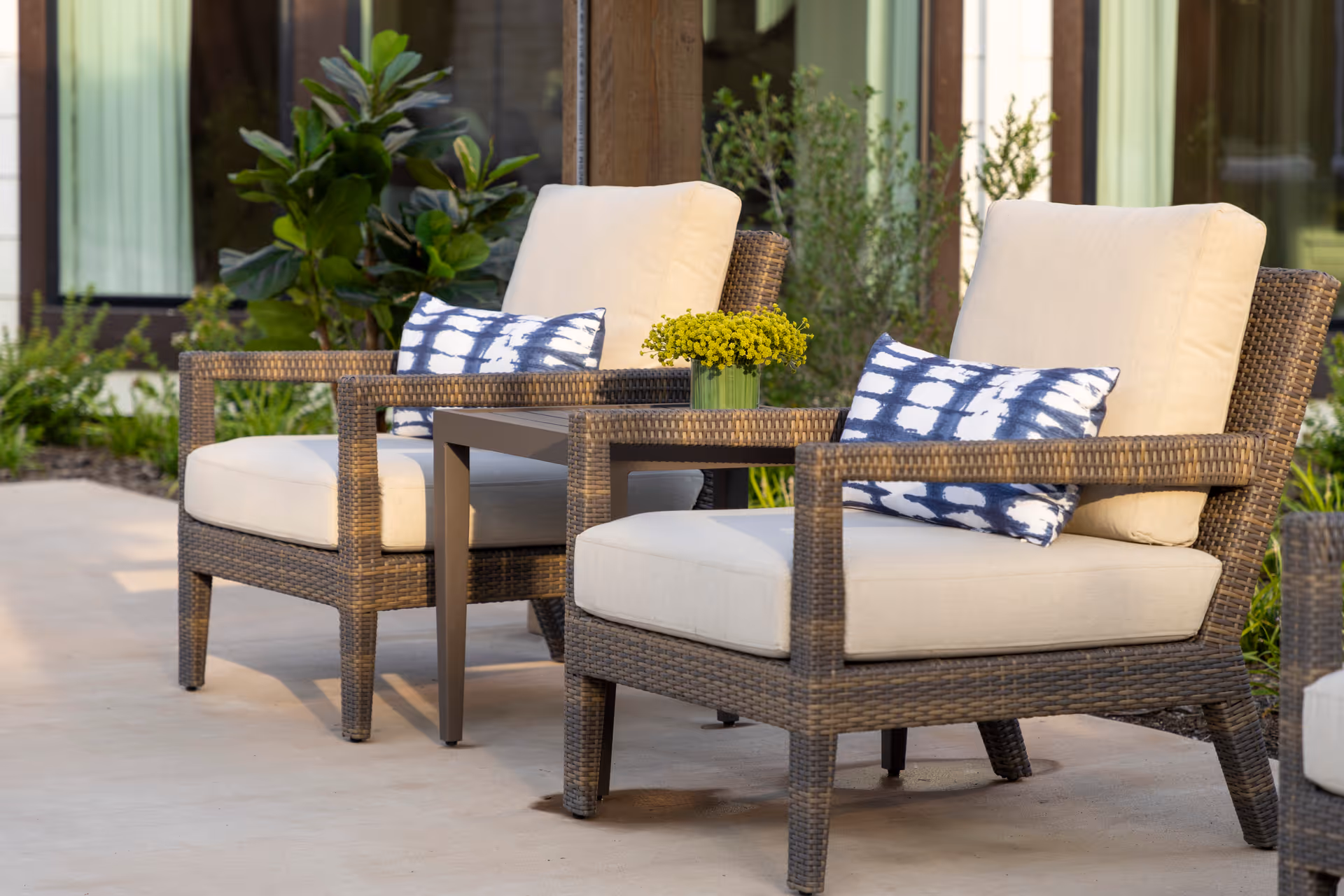 Two wicker patio chairs with cream cushions and blue patterned pillows flanking a small table with yellow flowers in an outdoor seating area.