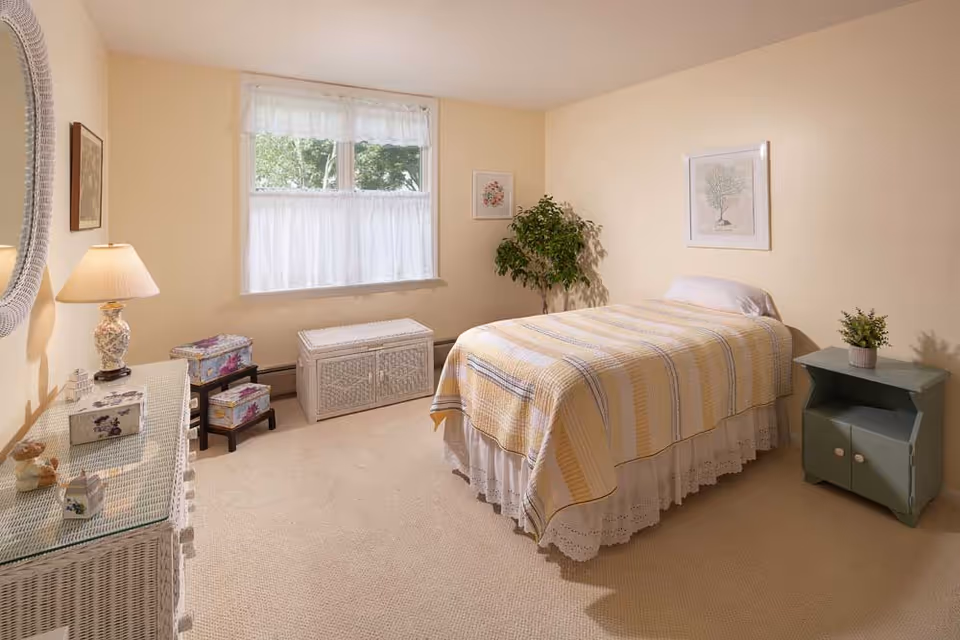 Bright, tidy bedroom with a single bed, wicker dresser, nightstand, lamp, and a window with curtains.