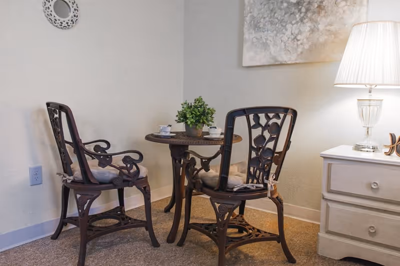 A cozy corner with two ornate wooden chairs with cushions around a small round table holding a potted plant and two teacups. Next to the table is a white nightstand with a glass lamp and a decorative wall mirror and painting above.