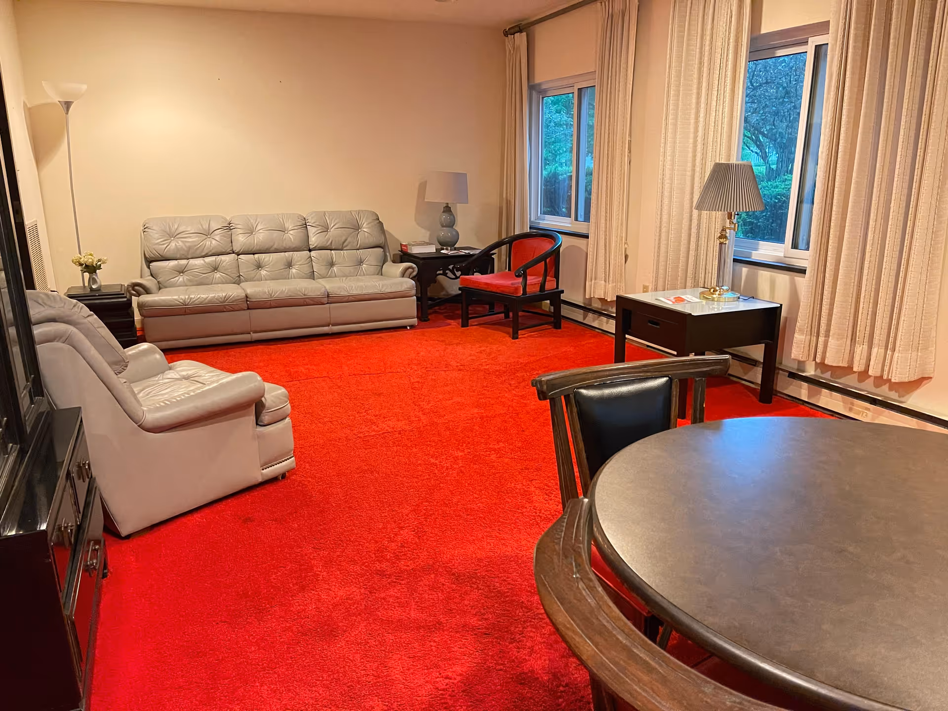 Living room with bright red carpet, gray leather sofas and chairs, a round table, side tables and windows with beige curtains.