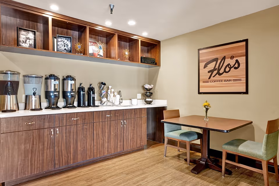Interior view of a coffee bar area named Flo's Coffee Bar in Terraza Court Senior Living, featuring a wooden counter with multiple beverage dispensers, coffee supplies, and a small table with two chairs and a small vase with flowers.