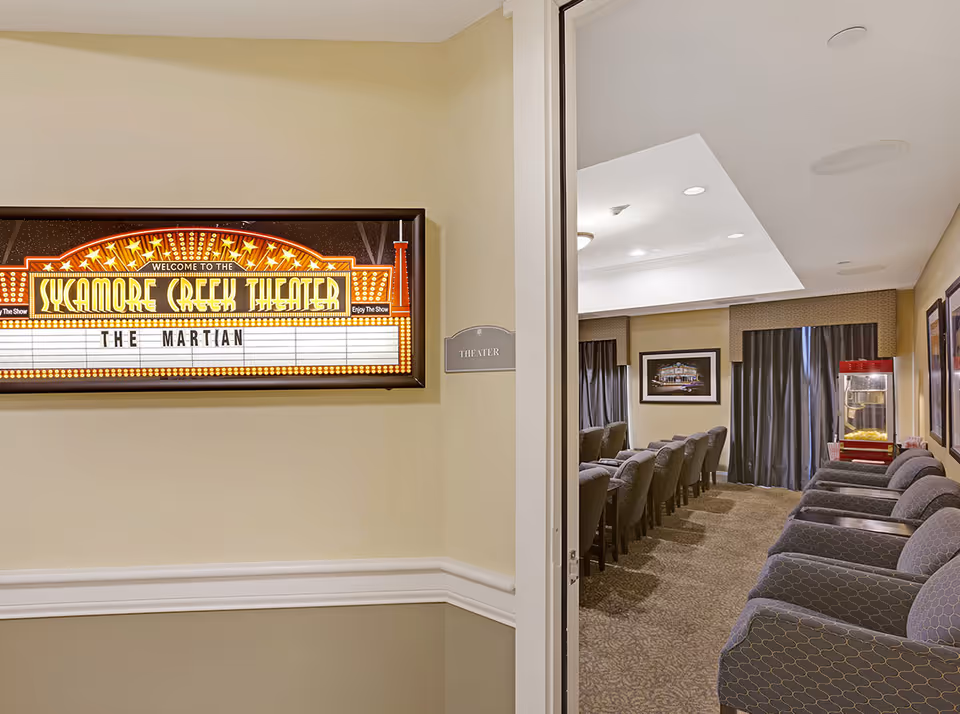 View into a small theater room with rows of cushioned chairs facing a screen. A popcorn machine is visible in the back corner. Outside the theater room, a wall sign reads 'THEATER' and a marquee sign says 'Welcome to the Sycamore Creek Theater' with the movie title 'The Martian' displayed.