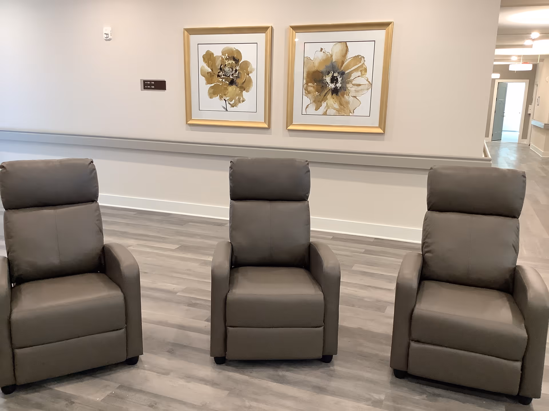 Three gray recliner chairs arranged in a row in a hallway with floral wall art and wood-look flooring.