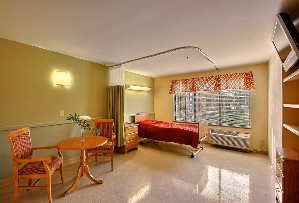 A well-lit room in a healthcare facility featuring a single hospital bed with a red blanket, a bedside table, two wooden chairs with red cushions, and a small round wooden table with a vase of flowers. There is a large window with blinds and a patterned valance, an air conditioning unit below the window, and a wall-mounted television on the right side. A privacy curtain is partially drawn around the bed.