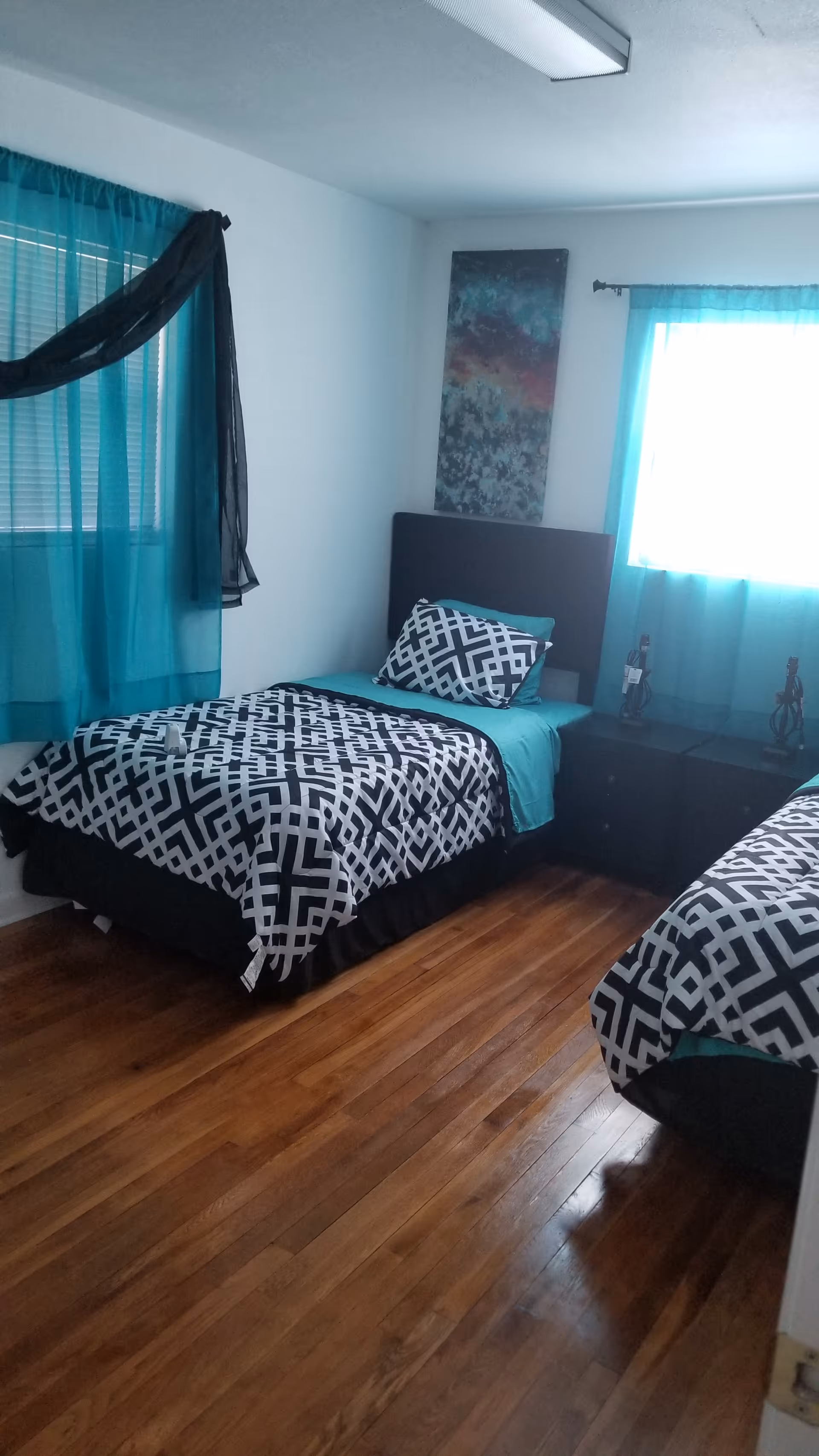 A bedroom with two single beds featuring black and white geometric patterned bedding and teal sheets. The room has wooden floors, two windows with teal curtains, a black nightstand between the beds, and a colorful abstract painting on the wall above one bed.