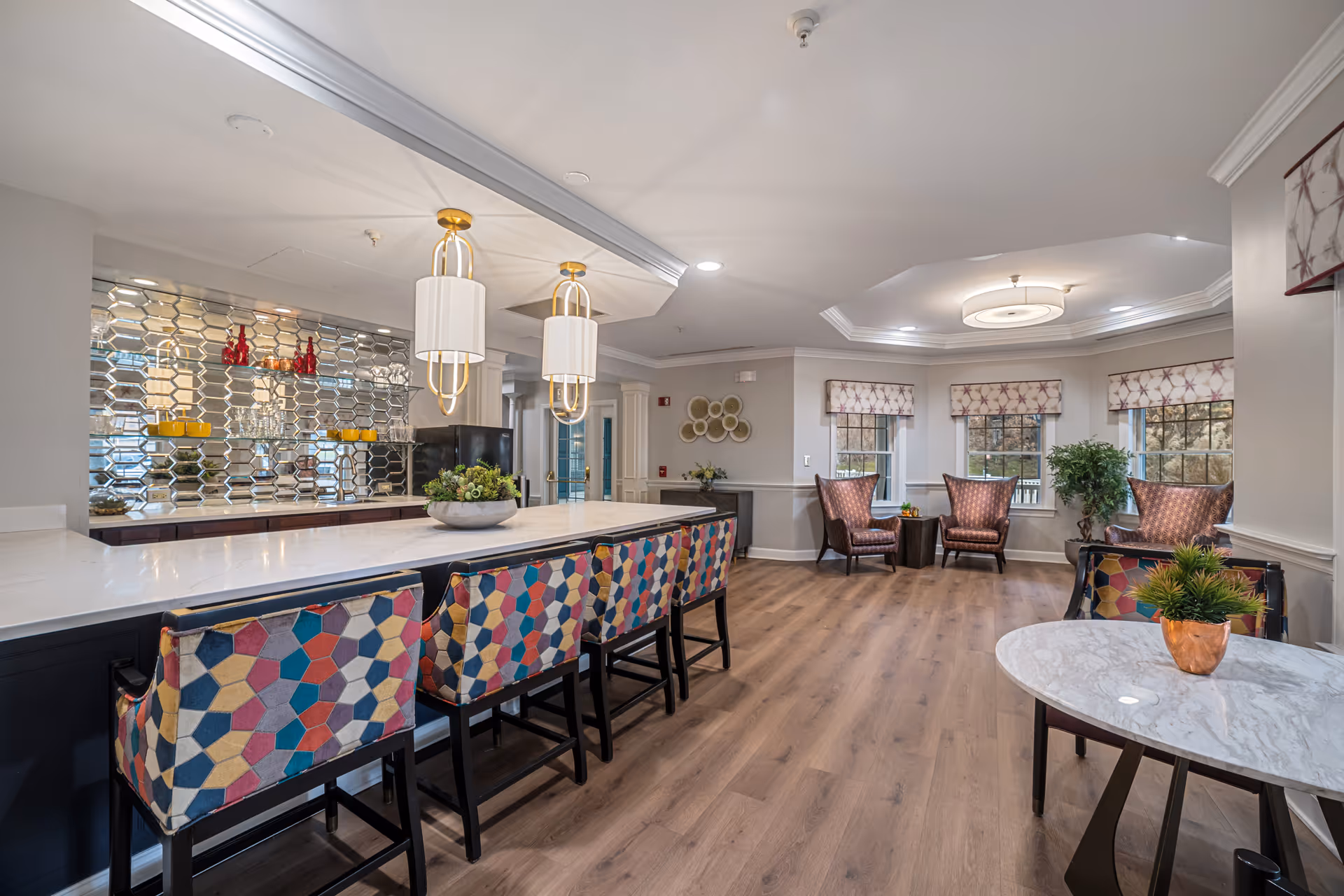 Open communal lounge and dining area with a marble-topped bar, colorful barstools, pendant lights, and seating by windows.