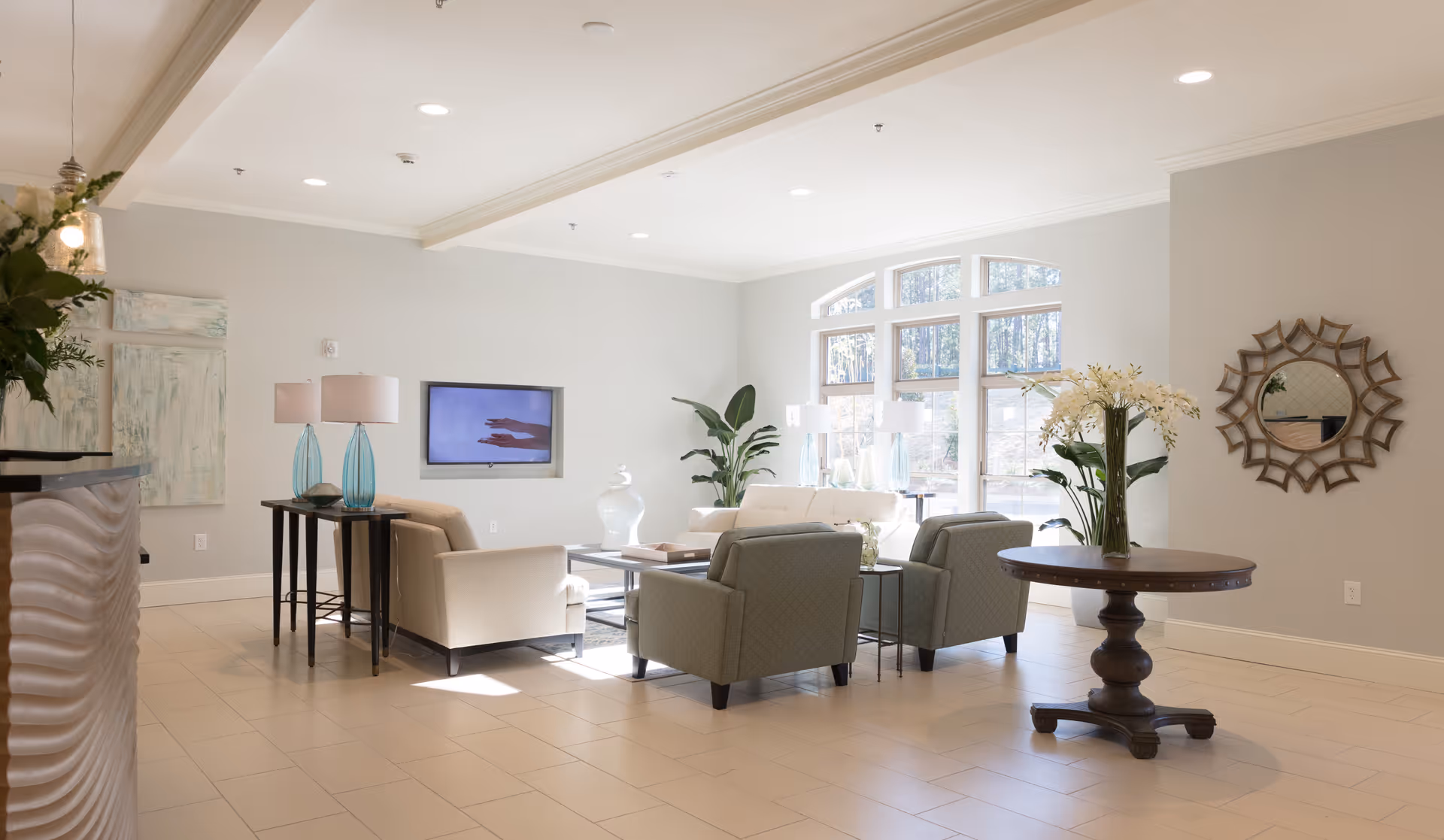 A bright and spacious living room area with large windows letting in natural light. The room features a white sofa, two green armchairs, a coffee table, and side tables with blue glass lamps. There is a round wooden table with a vase of flowers and a decorative round mirror on the wall. The walls are painted light gray and the floor is tiled in a light color.