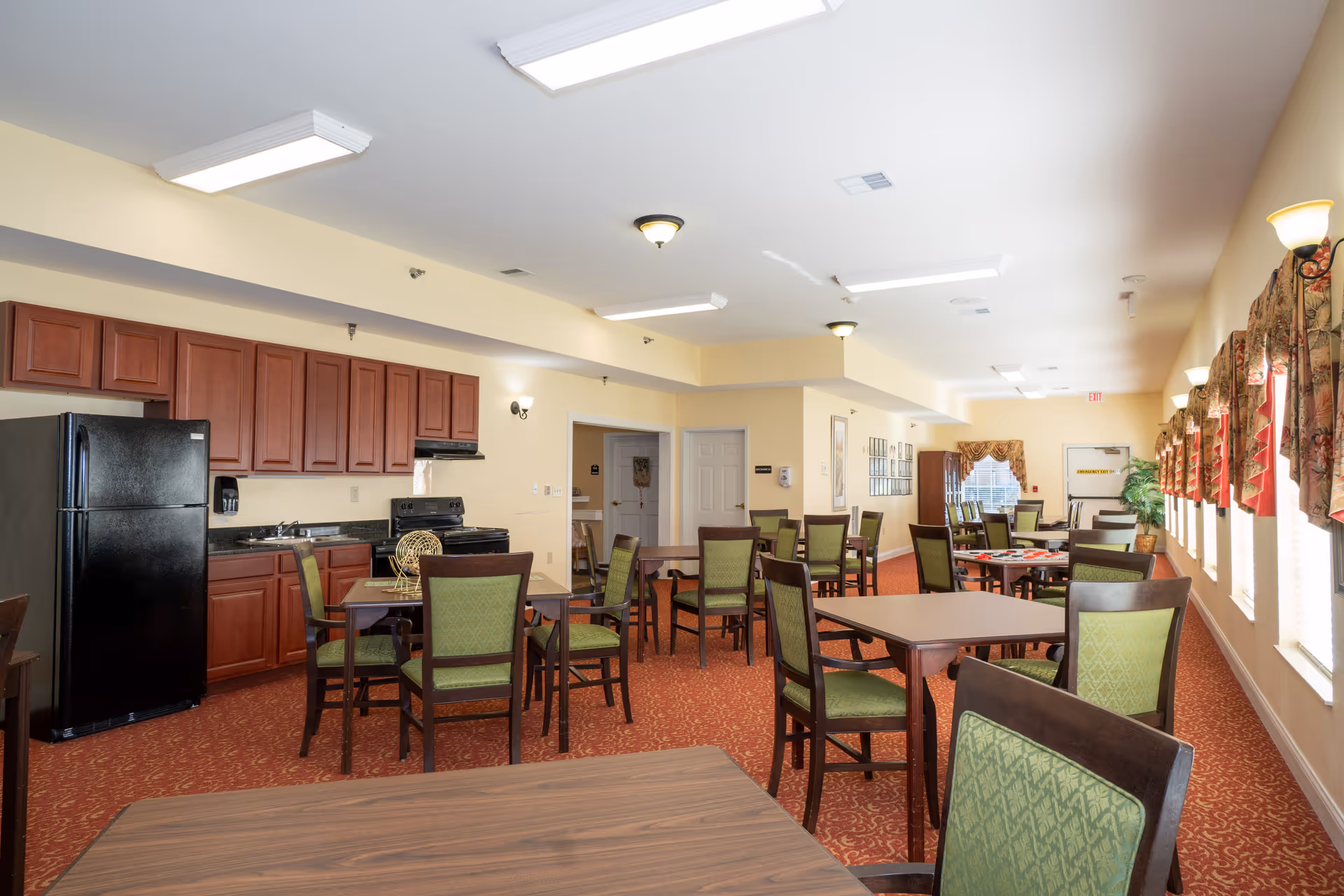A spacious dining room with multiple square tables and green cushioned chairs. The room has a red patterned carpet, beige walls, and several windows with floral valances letting in natural light. On the left side, there is a kitchen area with dark wood cabinets, a black refrigerator, stove, and sink. Ceiling lights and wall sconces illuminate the room.