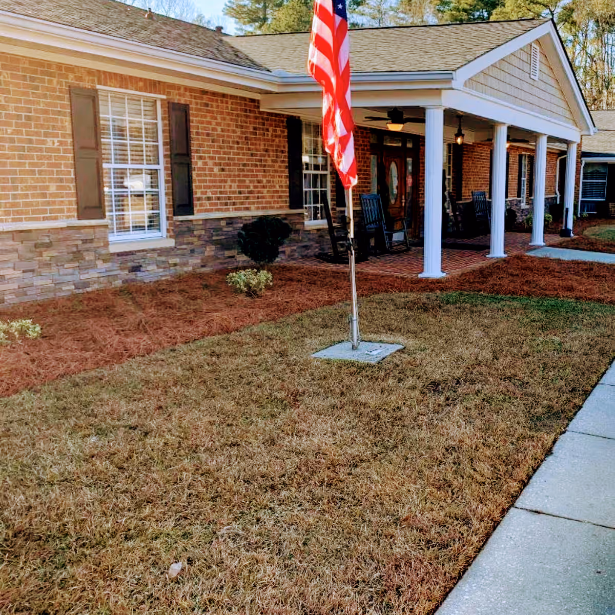Front exterior view of a brick building with a covered porch supported by white columns, rocking chairs on the porch, an American flag on a pole in the front yard, and a sidewalk leading to the entrance.
