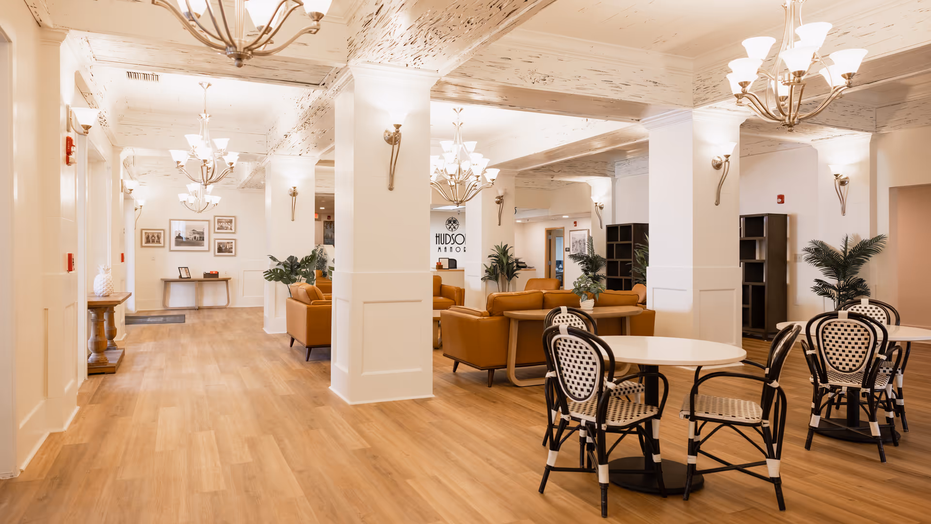 Spacious and well-lit interior of Hudson Manor Assisted Living featuring wooden flooring, white walls with decorative molding, multiple round tables with black and white chairs, several brown leather sofas, large white pillars, and elegant chandeliers hanging from a distressed white ceiling. There are framed pictures on the walls and green plants placed around the room.