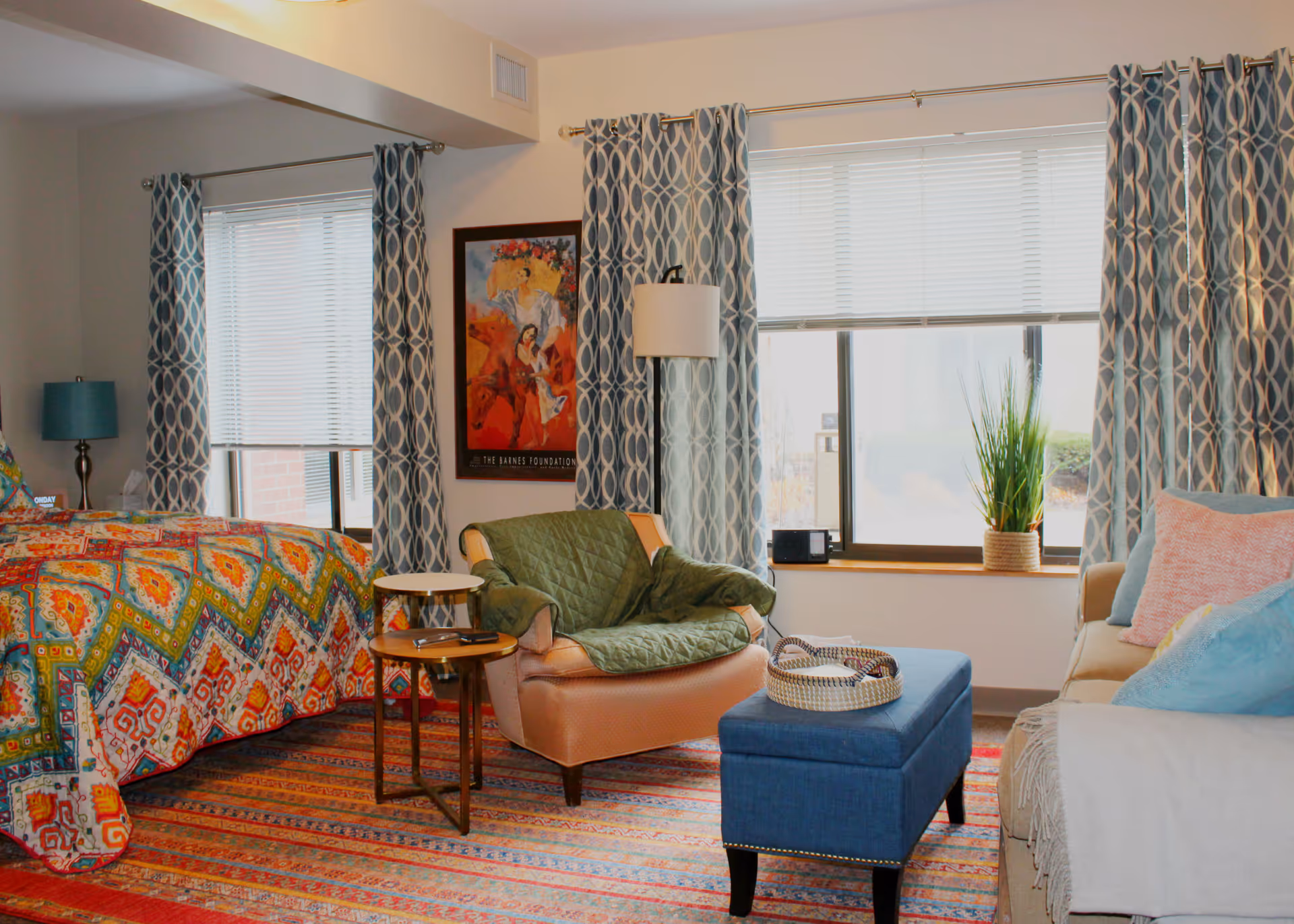 Bright senior living suite with a colorful bed, armchair, sofa, ottoman and patterned curtains beside a large window.