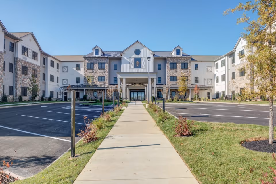 Front exterior view of Mercer Hill at Doylestown, a three-story senior living facility with a mix of stone and siding facade, a covered entrance, parking spaces on either side of a central walkway, and landscaped greenery.