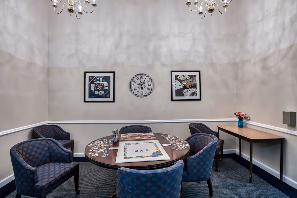 A small room with a round table in the center covered with a partially completed jigsaw puzzle and puzzle pieces scattered around. Six blue patterned chairs surround the table. On the wall behind the table are two framed pictures featuring playing cards and a clock in the middle. A small rectangular table with a blue vase holding pink flowers is against the right wall. The room has light-colored walls with a white chair rail and a dark carpeted floor. Two chandeliers hang from the ceiling.