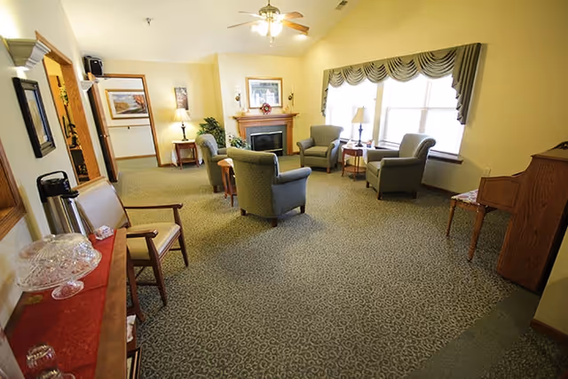 A cozy living room area with several upholstered armchairs arranged around a wooden fireplace. The room has a patterned carpet, large windows with valances, a ceiling fan with lights, and various lamps and framed artwork on the walls. There is a small wooden table and a cabinet along the walls.