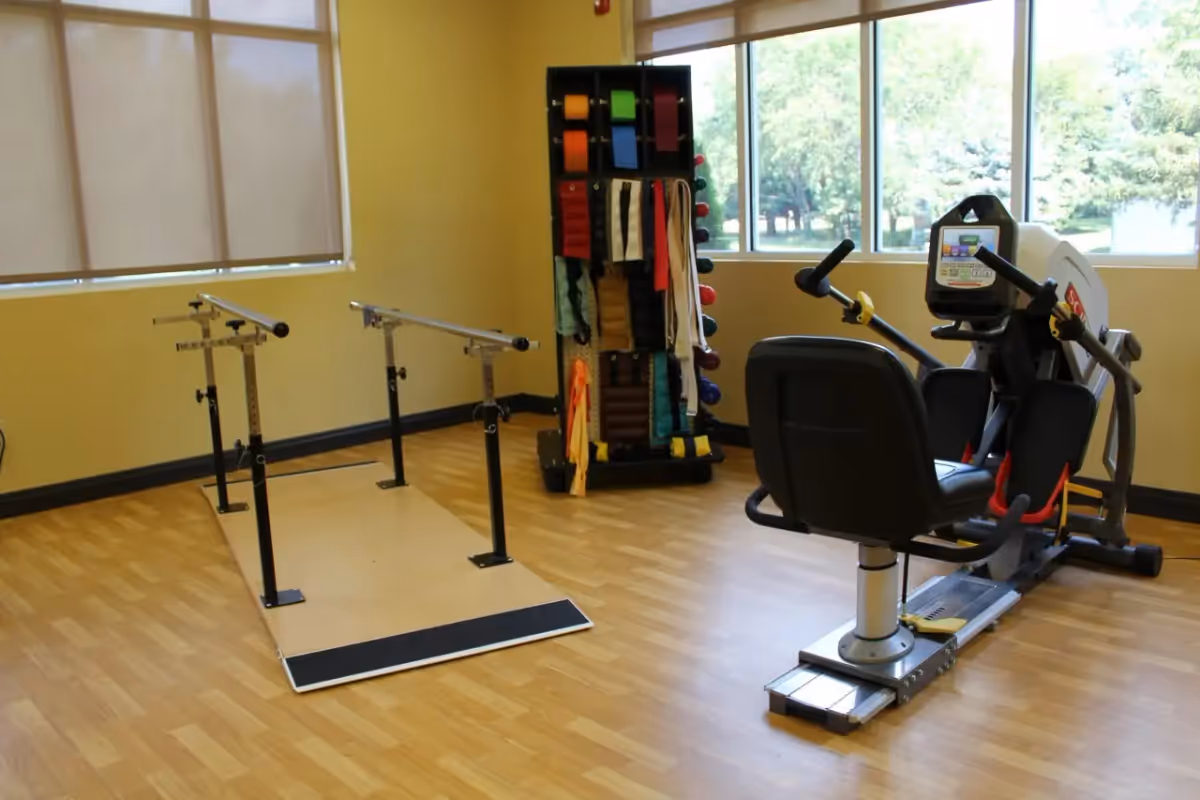 A physical therapy or exercise room with wooden flooring, featuring parallel bars for walking exercises, a recumbent exercise bike, and a rack holding various resistance bands and exercise equipment. Large windows provide natural light and a view of greenery outside.