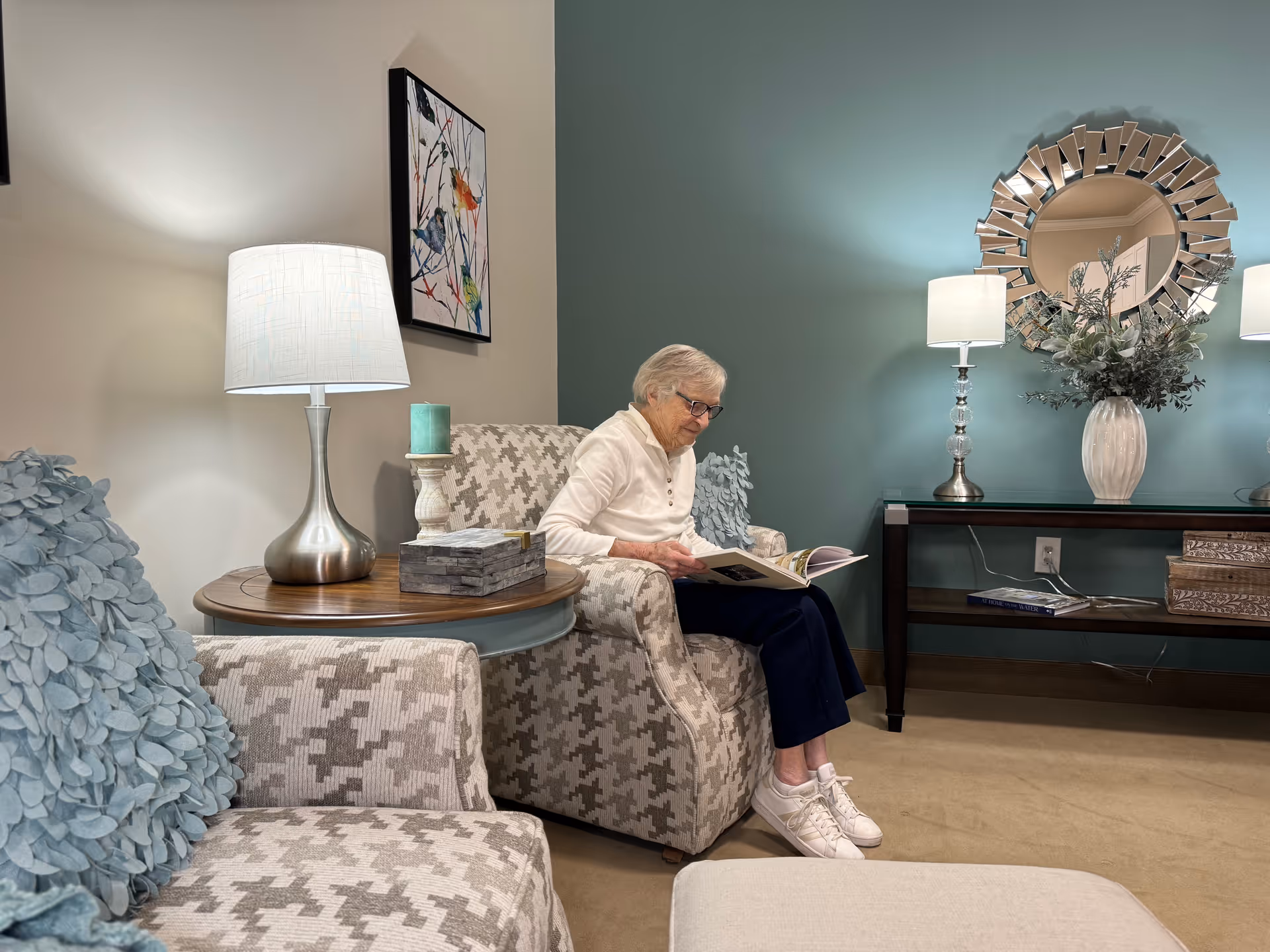 An elderly woman with white hair and glasses sits in a patterned armchair reading a book in a cozy living room. The room features a round wooden side table with a silver lamp and a green candle, a teal accent wall with a decorative round mirror, two table lamps, a vase with greenery, and a console table with books and decorative boxes. Another patterned armchair with a blue textured pillow is partially visible in the foreground.
