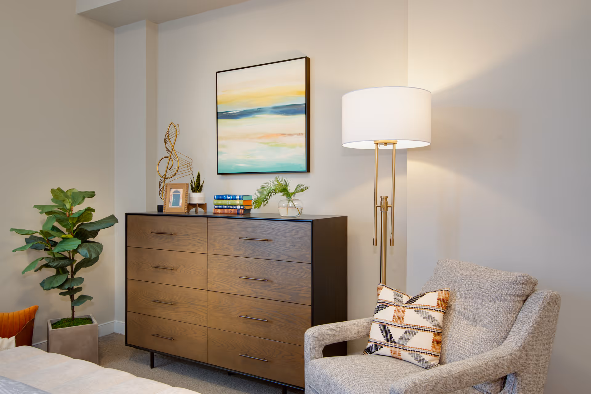 A cozy bedroom corner with a wooden dresser, potted plant, floor lamp, armchair with a patterned pillow, and framed artwork above the dresser.