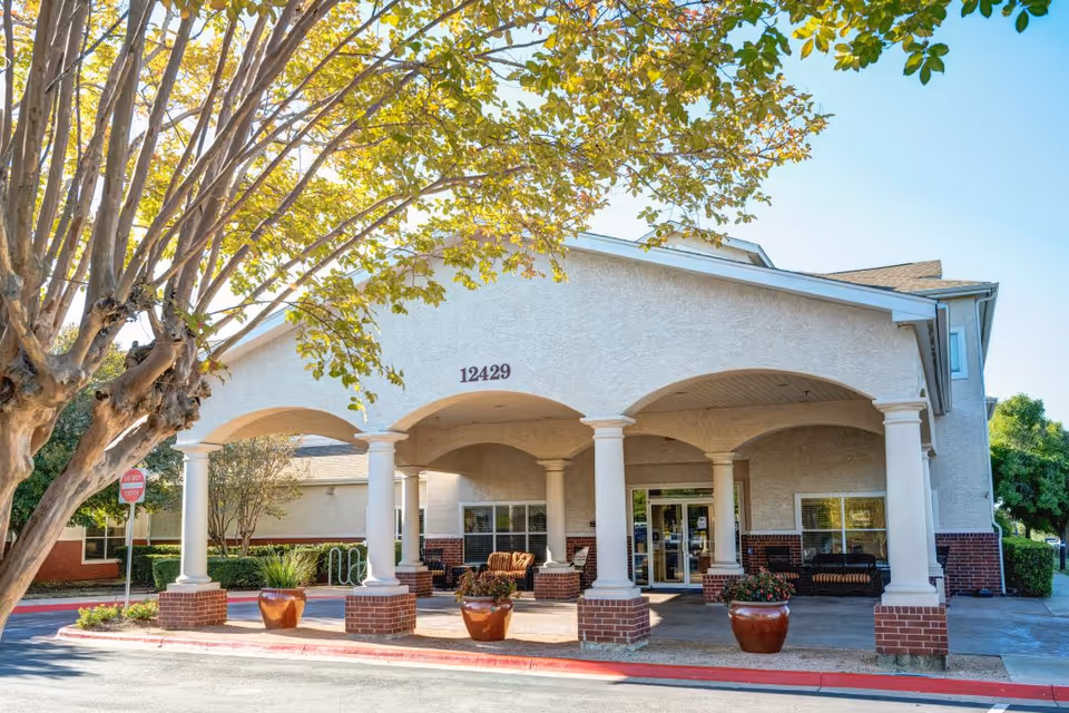 Front exterior view of a senior living facility building with the address number 12429 displayed above the entrance. The entrance features a covered portico supported by white columns with brick bases. There are large potted plants and outdoor seating visible near the entrance. A tree with green leaves is in the foreground on the left side.