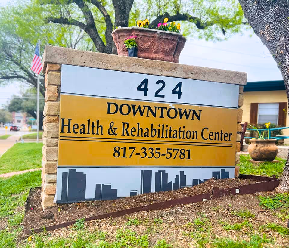 Outdoor stone sign for Downtown Health & Rehabilitation Center with the address number 424 and phone number 817-335-5781, surrounded by grass, trees, and a flower pot on top of the sign.