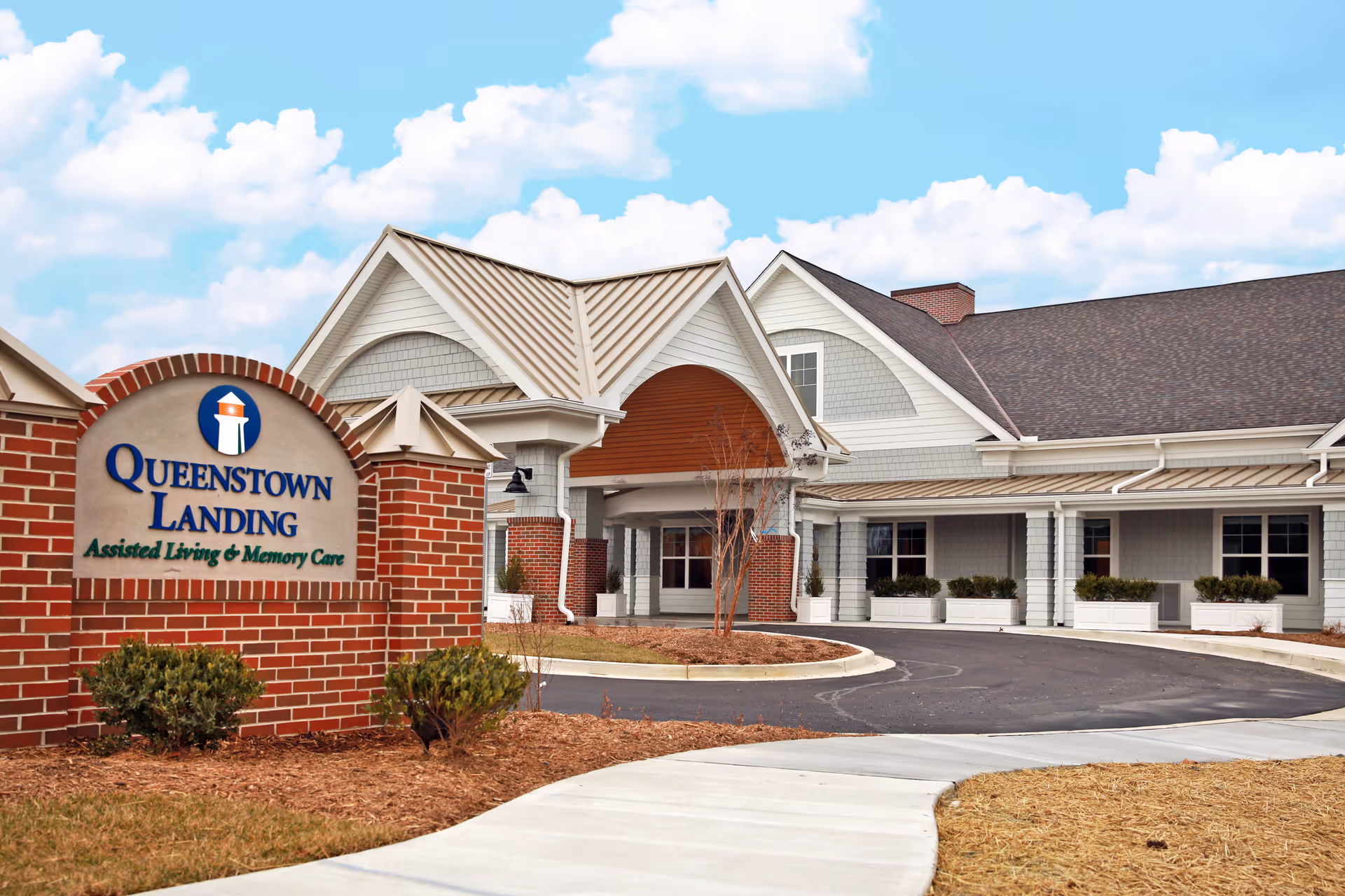 Exterior view of Queenstown Landing, an assisted living and memory care facility, showing the building entrance with a covered driveway, brick and siding facade, and a sign with the facility name in front.