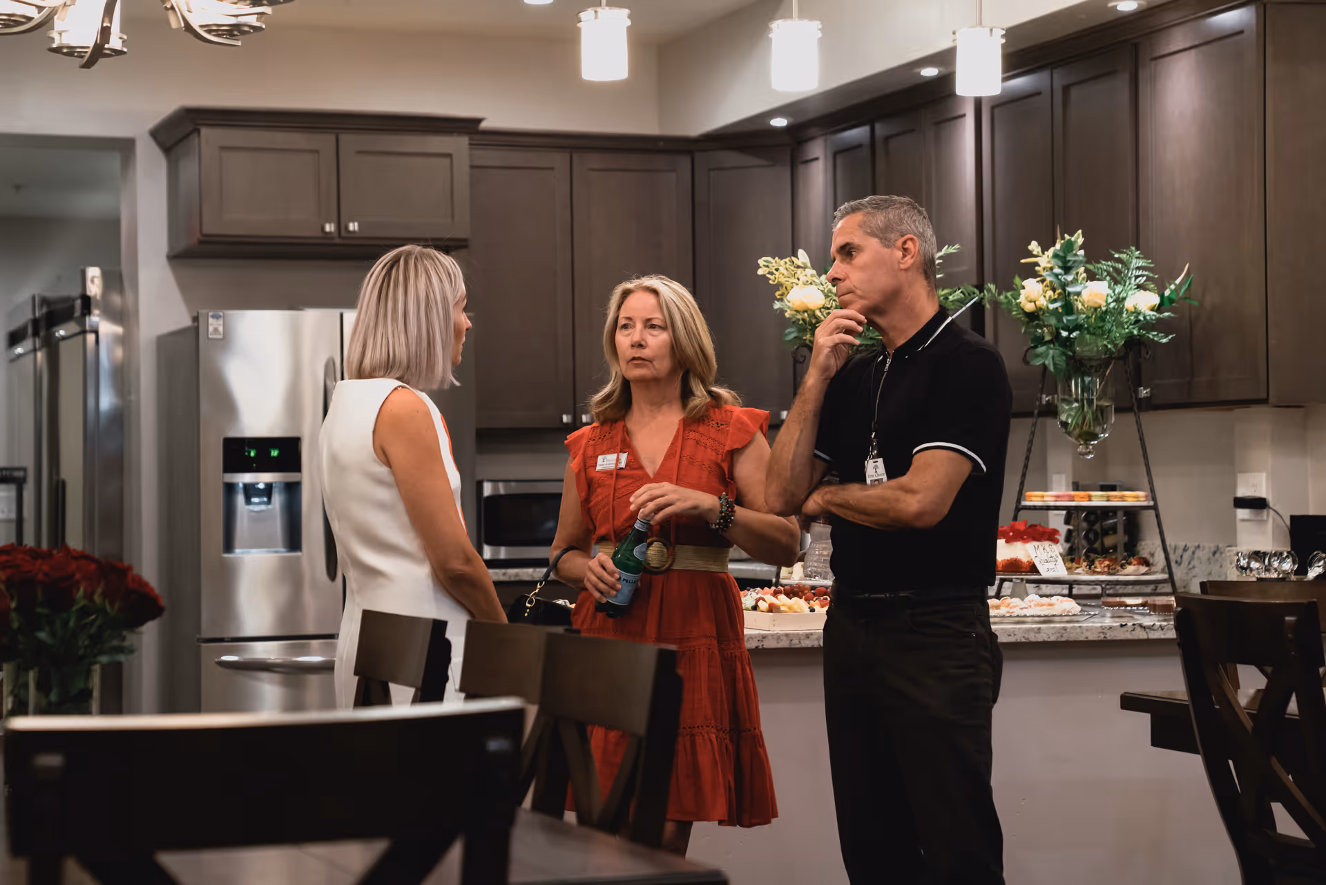 Three adults engaged in conversation in a modern kitchen with dark wood cabinets, stainless steel refrigerator, and a countertop with flowers and assorted food items.
