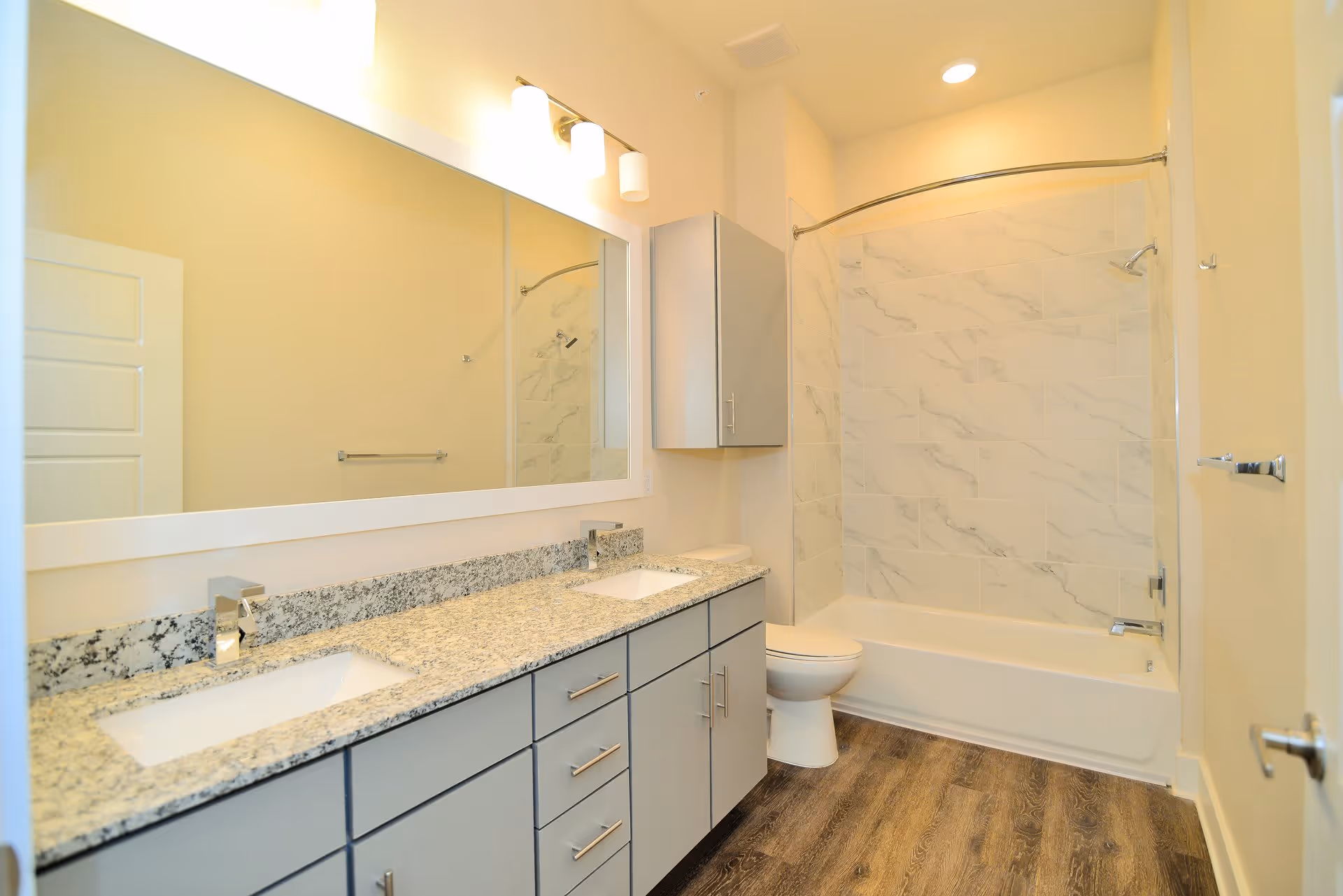 A modern bathroom featuring a double sink vanity with granite countertop and gray cabinets, a large rectangular mirror above the sinks, a toilet, and a bathtub with a curved shower curtain rod and marble-patterned tile surround. The floor has wood-like vinyl flooring and the walls are painted light beige.
