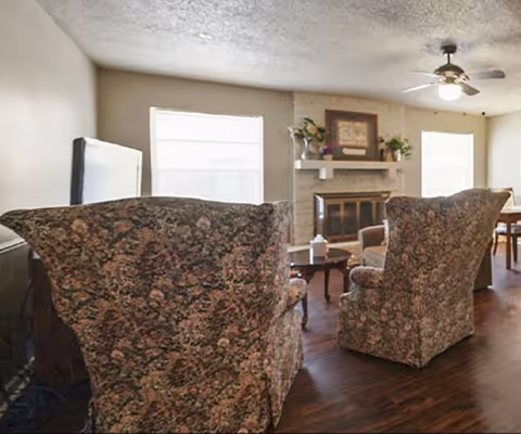 A living room with two floral upholstered armchairs facing a fireplace. The room has wooden flooring, a ceiling fan with a light, and two windows letting in natural light. There is a TV on the left side and a small table between the chairs with a decorative item on it.