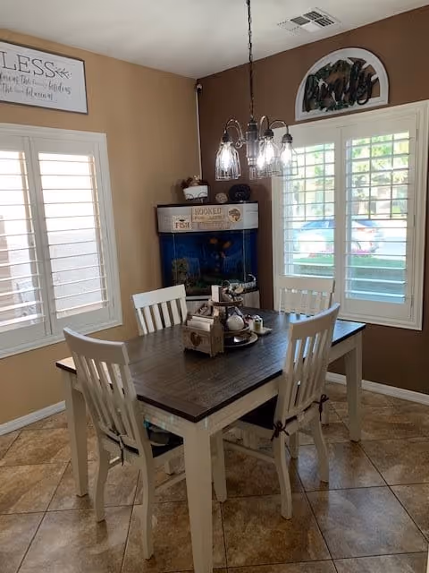 A dining area with a rectangular wooden table and four white chairs. The table has decorative items including a small wooden box and a tray with various objects. There is a fish tank in the corner against the wall, and two windows with white shutters letting in natural light. Above the windows are decorative wall hangings, and a chandelier with five lights hangs above the table. The walls are painted brown and the floor is tiled.