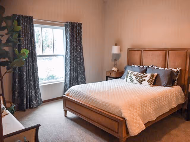 A cozy bedroom with a wooden bed frame, white quilted bedspread, and decorative pillows. There is a nightstand with a lamp beside the bed, a window with patterned curtains, and a potted plant in the corner.