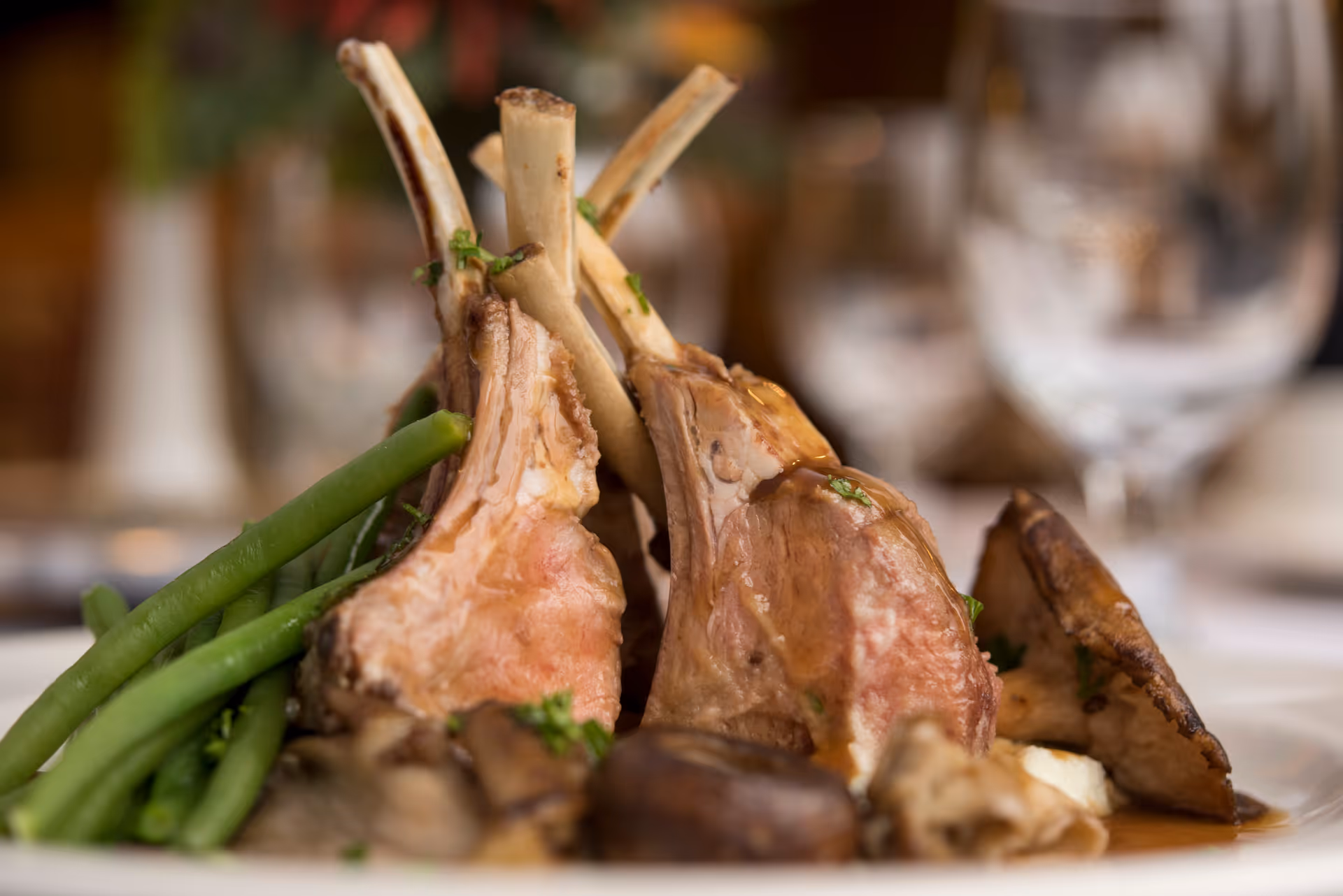 Rack of lamb chops served with green beans and mushrooms on a plate with blurred wine glasses in the background.