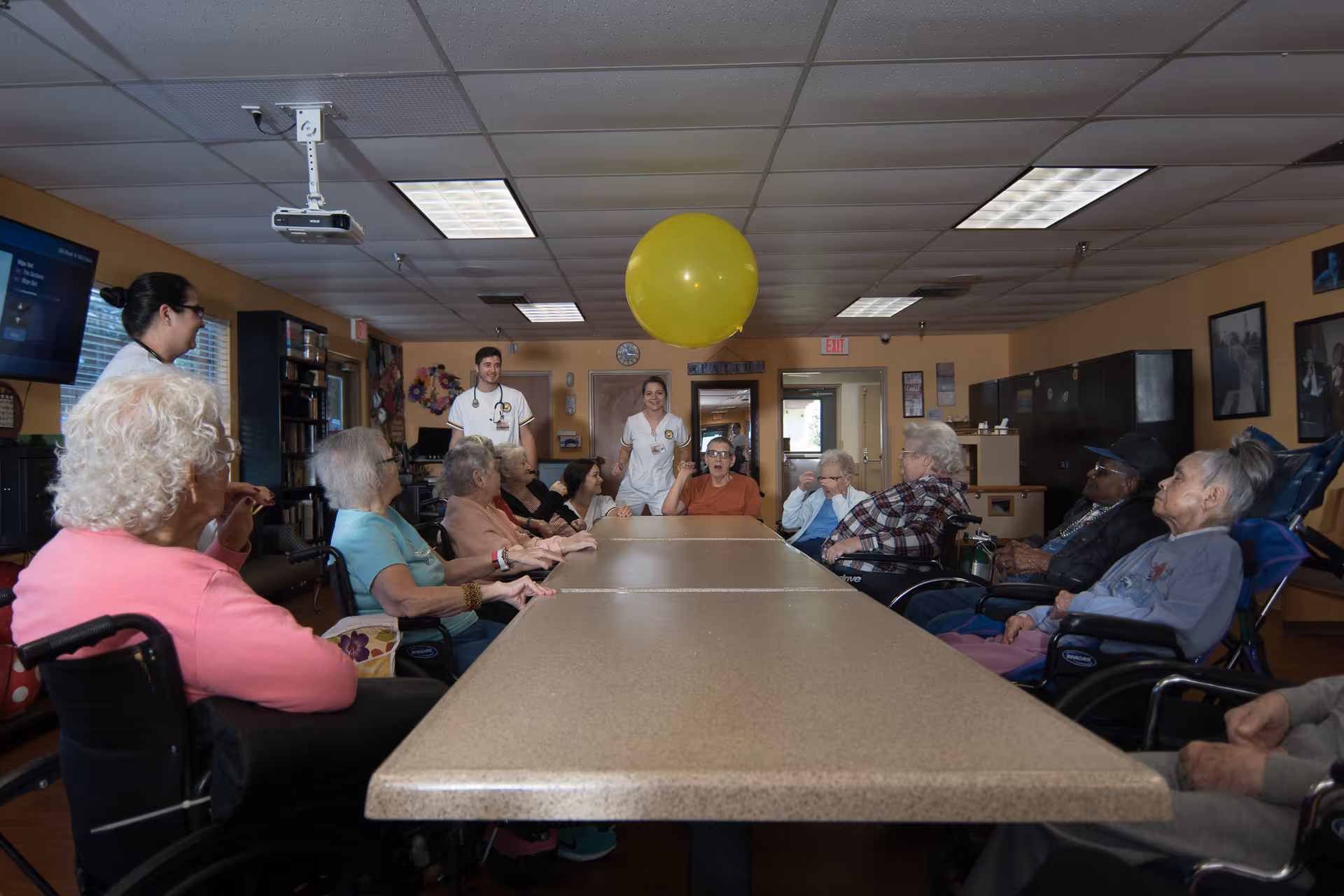 Elderly residents and staff sit around a long table in a common room playing with a large yellow balloon.