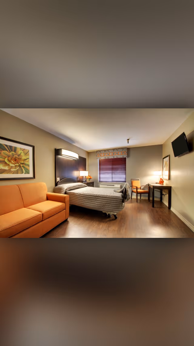 Furnished senior living bedroom with a bed, orange sofa, desk and wall-mounted TV facing a window with blinds.