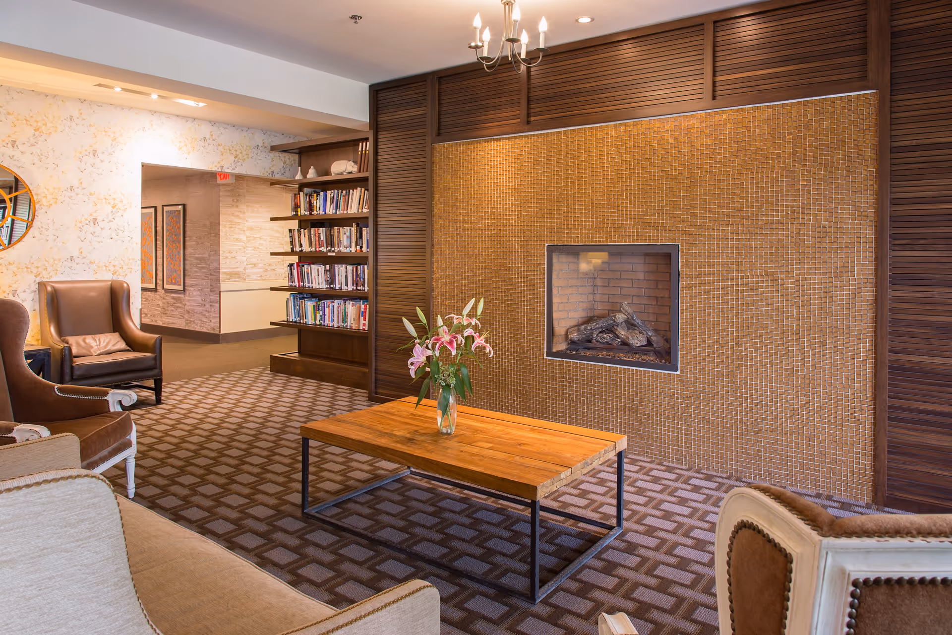 A cozy living room area with a wooden coffee table holding a vase of pink lilies, surrounded by upholstered chairs and a sofa. The room features a fireplace with a mosaic tile surround and a built-in bookshelf filled with books. The carpet has a geometric pattern and the walls have a textured design.