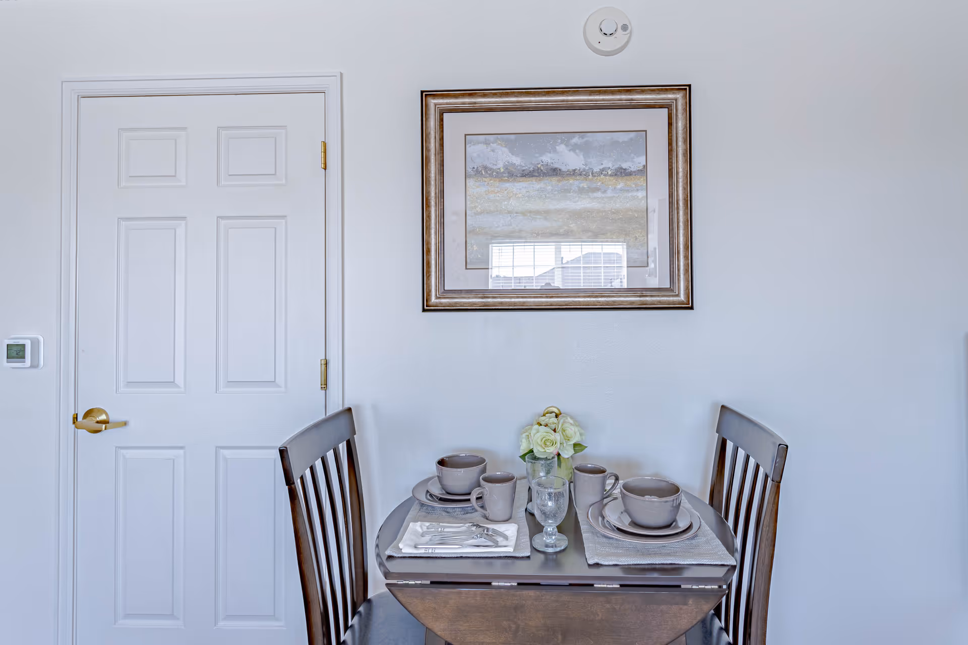 A small dining area with a round wooden table set for two, featuring beige plates, bowls, mugs, and silverware on placemats. A small vase with white flowers is centered on the table. Behind the table is a white door with a gold handle and a framed abstract painting on the wall.