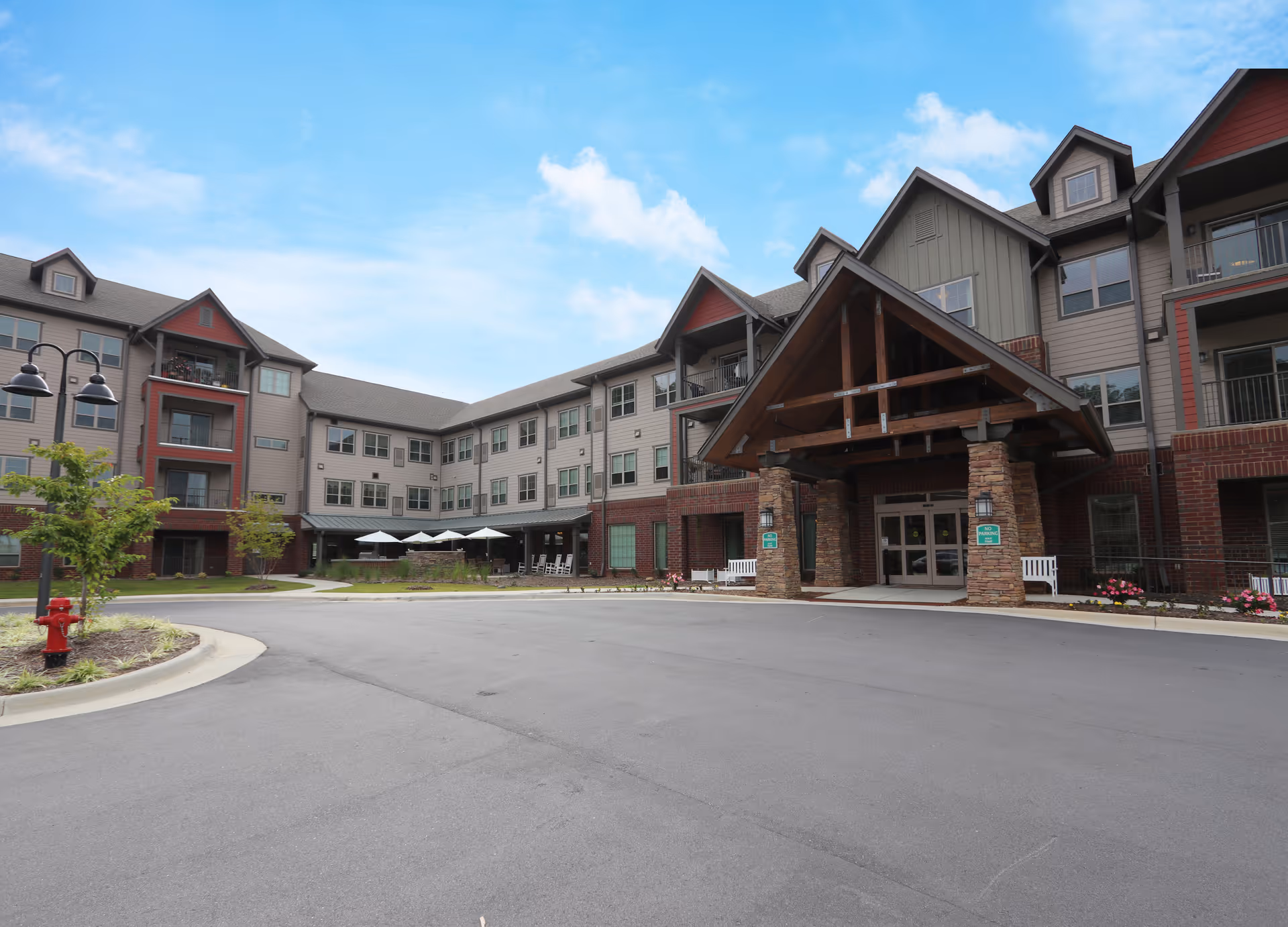 Front entrance of a multi-story senior living building with a covered porte-cochère, driveway and outdoor seating with umbrellas.