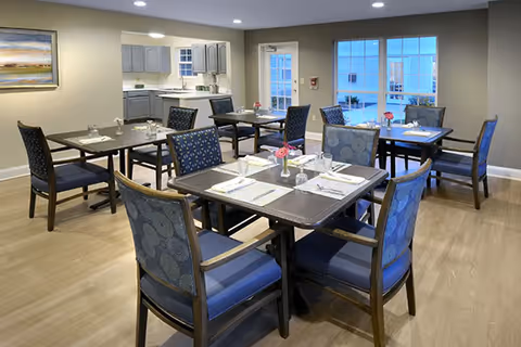 A dining room in a senior living facility with several square tables set with placemats, napkins, glasses, and small flower vases. The room has wooden flooring, cushioned chairs with patterned upholstery, and a kitchen area visible in the background. Large windows and a glass door let in natural light.
