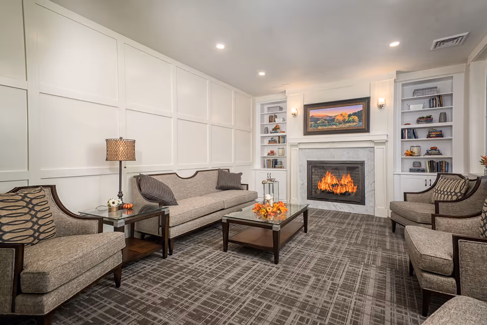 A cozy senior living facility lounge with a lit fireplace, a beige sofa, two armchairs, a glass coffee table with autumn decorations, built-in white bookshelves, and a framed landscape painting above the fireplace.