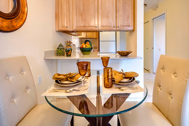 A small dining area with a round glass table set for two, featuring beige cushioned chairs, amber-colored glassware, and gold-toned napkins. Behind the table is a kitchen counter with wooden cabinets, a refrigerator, and decorative items including a bowl of fruit and jars. The walls are painted light yellow, and a round decorative mirror is partially visible on the left wall.