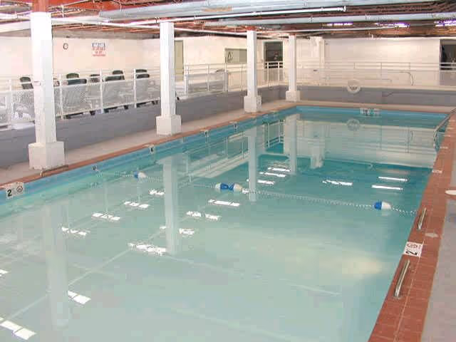 Indoor swimming pool with clear water, surrounded by a tiled deck and white railings. There are several white support columns and a row of chairs along one side of the pool area.