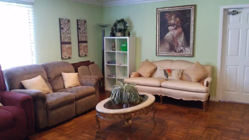 A cozy living room with a gray recliner sofa and a beige sofa with cushions. There is a glass-top coffee table with a potted plant in the center. A white shelving unit with decorative items and a floor lamp are against a light green wall. Two framed artworks hang on the walls, and a closed white door is visible on the right side.