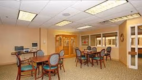 Interior senior living common area with round tables, teal chairs, and computer workstation.
