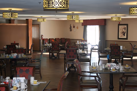 A dining room in an assisted living facility with multiple tables and chairs arranged neatly. The tables have place settings including cups, plates, and napkins. The room features wooden flooring, decorative ceiling lights, and wall art. There is a window with curtains allowing natural light into the room.