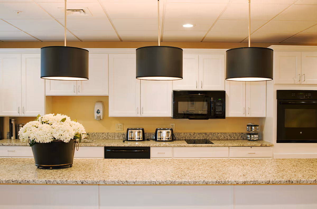 A modern kitchen area with white cabinets, granite countertops, and three large black pendant lights hanging from the ceiling. On the countertop, there is a black pot filled with white flowers. The kitchen features two toasters, a microwave, an oven, and a stovetop. The walls are painted a light yellow color.