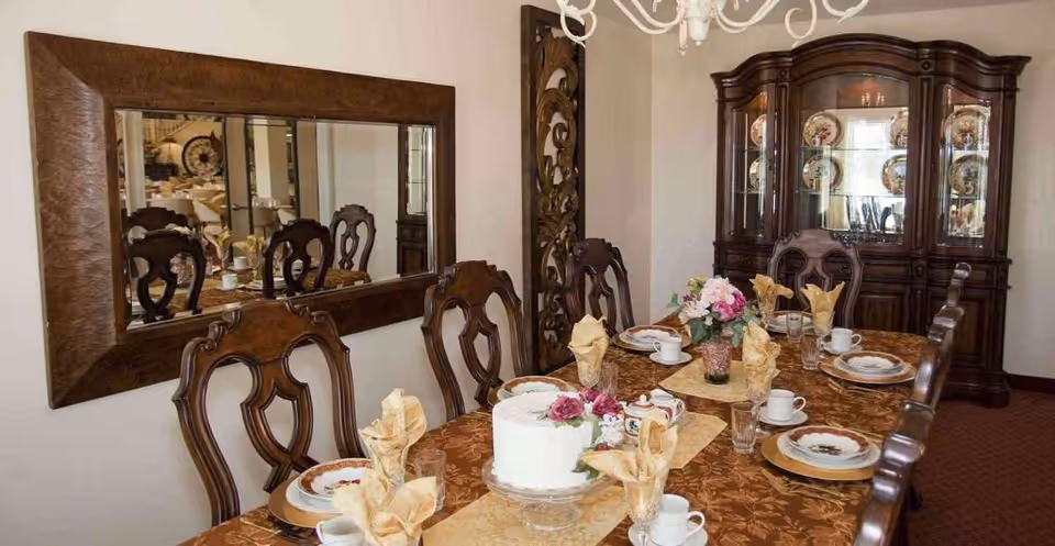 A formal dining room with a long wooden table set for a meal. The table is covered with a brown patterned tablecloth and decorated with a white cake, floral centerpiece, and neatly folded napkins. There are eight ornate wooden chairs around the table. A large wooden china cabinet with decorative plates and glassware is against the wall, and a large rectangular mirror hangs on the opposite wall reflecting the room.